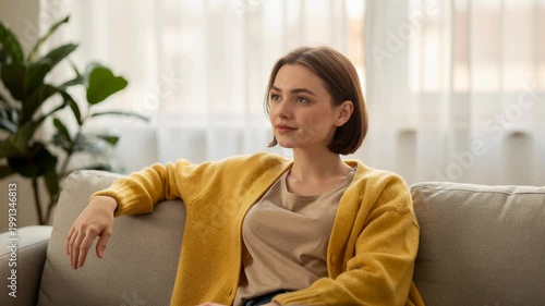 Young woman in cozy yellow cardigan relaxing thoughtfully on soft sofa at home