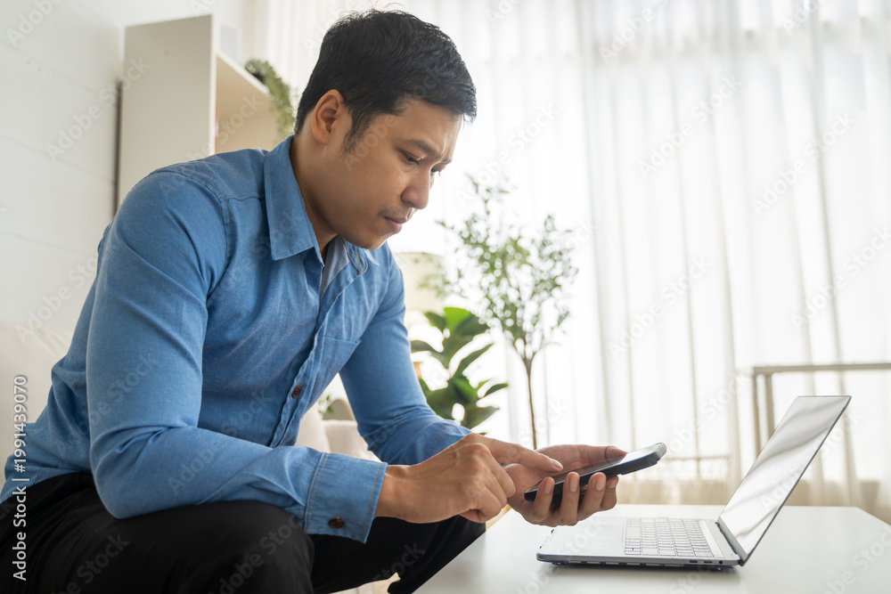 Fototapeta premium Serious asian businessman using smartphone and laptop on desk at home, male freelancer checking mobile banking app or trading online stocks, Remote working and financial investment concept