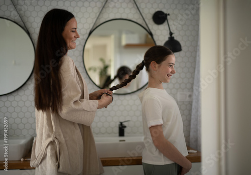 фотографія Mother braiding daughters thick hair in bathroom.
