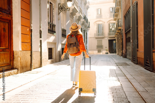 Fotografie A woman walks along a narrow street, pulling a bright yellow suitcase
