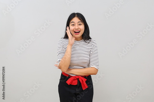 Foto Young Asian woman with surprised facial expression, receiving good news or surprise, mouth open in amazement, putting hands beside mouth and looking at camera