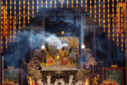 A central incense burner releases thick blue smoke within a traditional Chinese temple. Ornate gold decorations and peacock feathers adorn the burner while red and gold lanterns hang from above.