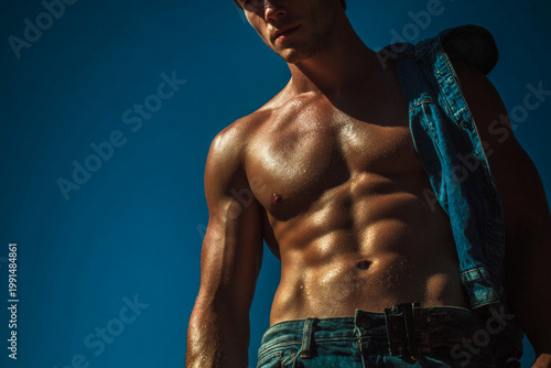 A handsome shirtless bodybuilder athlete displays a strong, muscular male torso with defined abs and chest muscles in a gym fitness setting