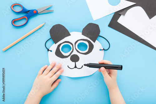 Little child boy hand drawing white black panda bear paper mask with marker on light blue table background. Pastel color. Point of view shot. Closeup. Creating decoration. Top down view.