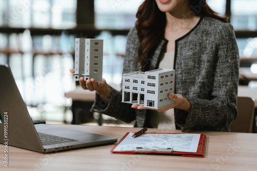 Businesswoman holding a small building model at a desk with a laptop and documents, representing real estate investment, property management, and financial decision-making.