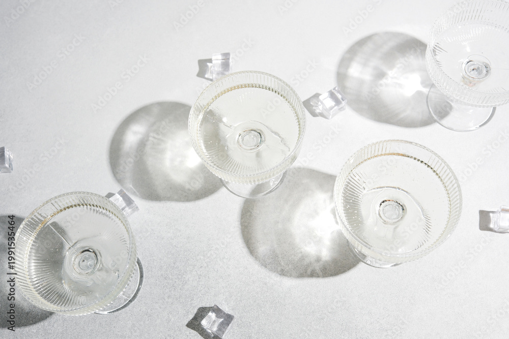 Obraz premium Four coupe glasses with clear liquid and ice cubes on a textured white surface creating strong shadows.