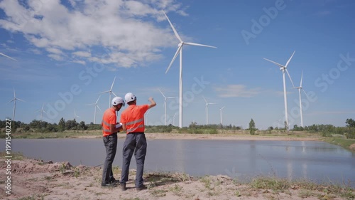 Rear view of Asian engineers pointing at wind turbines during inspection at a renewable energy site, representing clean energy operations and teamwork in sustainable infrastructure.