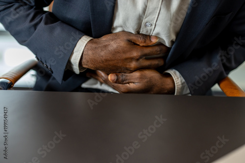 Fotografie Businessman in suit seated at desk clutching his abdomen in visible discomfort,