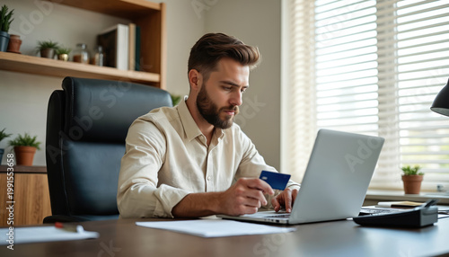 Bearded man pays online using credit card and laptop at desk. Person works in office with computer and card. Manager completes purchase or transaction.