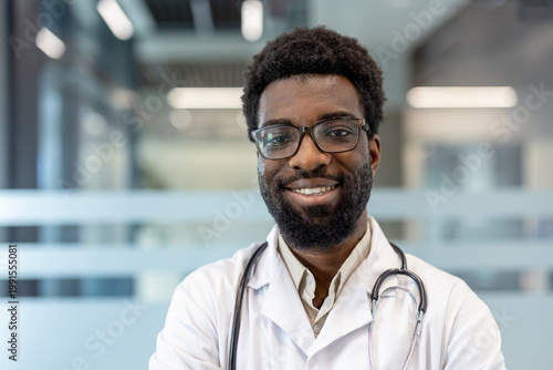 Male professional doctor looking at camera and smiling, wearing a white lab coat and stethoscope, posing against a modern clinic background, representing healthcare expertise and patient care