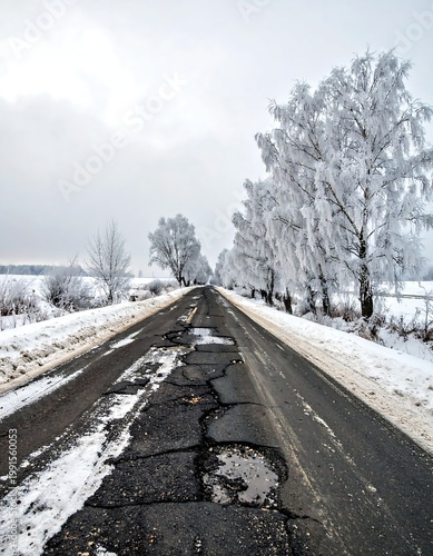 Damaged Asphalt Road With Deep Potholes And Cracks Winding Through A Winter Landscape With Frost Covered Trees Under An Overcast Grey