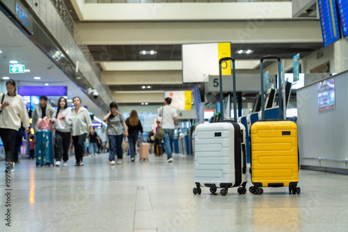 Two modern hard-shell suitcases, yellow and white, are standing in a blurred airport terminal departure hall near check-in counters and flight information displays, ready for travel and vacation.