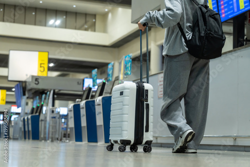 Close-up of a traveler's lower body standing and holding a white suitcase with an extendable handle in a busy airport terminal near check-in counters and baggage claim, ready for travel.