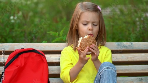 Schoolchild sits on bench with backpack in schoolyard and eats hamburger. Little girl has lunch outdoors. Education school