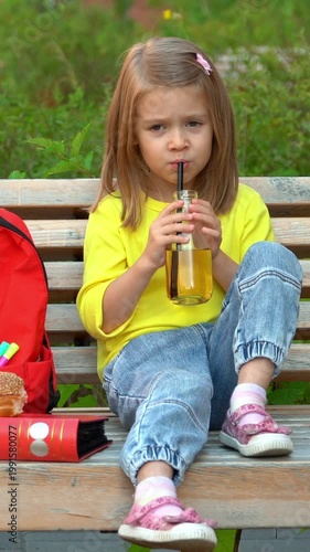 Little schoolchild with backpack eats school lunch and drinks juice outdoors. Education concept. Vertical shot