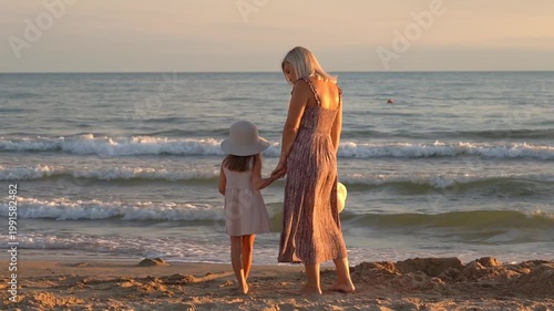 Back view of mother and daughter look at sea on sand beach at sunset. Happy family walk by ocean. Summer vacation travel holiday