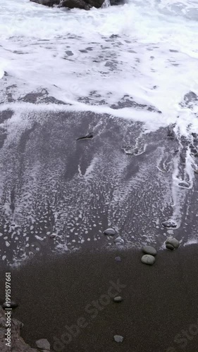 Close-up of white ocean foam washing over black volcanic sand with small pebbles on beach in Tenerife, Canary Islands. Perfect for nature, background and abstract themed projects