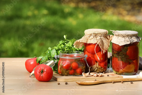 фотографія Jars of canned tomatoes with wooden board, spoon and parsley on table outdoors
