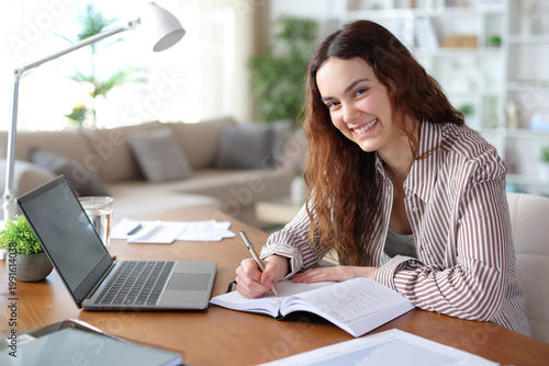 Happy teleworker writing in agenda looking at you working at home