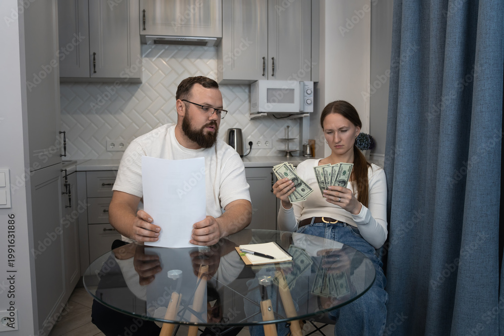 Fototapeta premium Couple reviewing financial documents and counting cash, facing budget challenges and money stress while dealing with household expenses in their modern kitchen