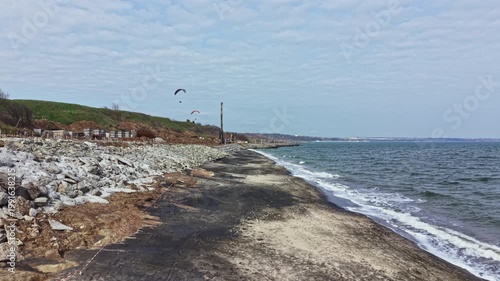 Kite surfers glide across the water as waves lap at the shore. A rocky beach stretches alongside grassy land under a cloudy sky at the coast.