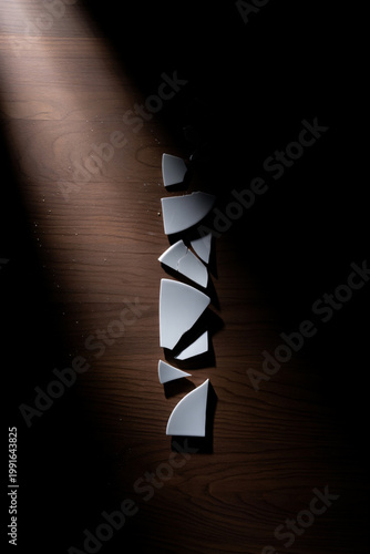 Broken porcelain plate on dark wooden floor with sharp shards, dramatic side lighting, minimalist metaphor for divorce or stress