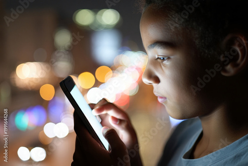 Close-up of teenager hands holding smartphone in dark room, screen light reflecting on face with bokeh city lights background