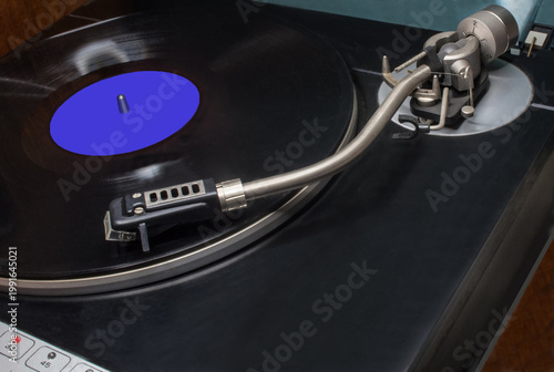 A vintage vinyl record player with a record mounted on it. Platter holding the record, the tonearm with the cartridge and stylus. Vinyl record with an unmarked blue sticker. 33 revolutions per min.	