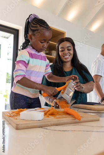African American mom and child grating carrot on board in kitchen, mom steadying grater over bowl