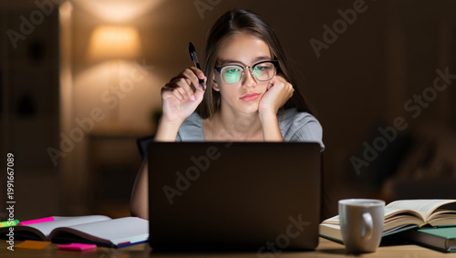 Tired teenage girl studying at desk at night with laptop and textbooks, focused expression under warm desk lamp light, cinematic student life