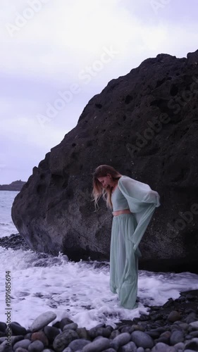 A vertical full-length shot of a joyful blonde woman in a flowing mint-green outfit laughing as sea foam washes over her feet next to a massive dark volcanic rock