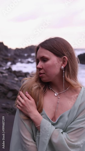 A vertical vertical shot of a charming blonde woman in a flowing mint-green outfit standing on a black sand beach, touching her hair, and wearing elegant pearl and silver earrings