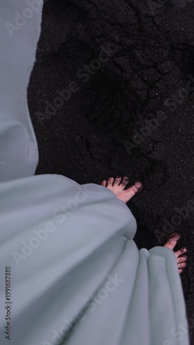 A cinematic vertical POV shot looking down at bare female feet and the edge of a flowing mint-green dress taking steps on dark, textured volcanic sand, with blurred ocean waves at the background