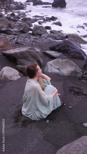 Vertical side view of a pensive woman in a flowing mint-green outfit sitting on a black volcanic sand beach surrounded by dark rocks, with ocean waves in the background