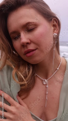 Vertical close-up of a smiling blonde woman looking at the camera, wearing long silver chain and pearl earrings, a matching necklace, and a mint-green top, against a soft blurred background of the oce