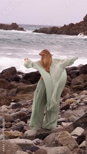 Vertical back view of a woman with long hair wearing a flowing mint-green outfit, standing on dark coastal rocks and looking at the ocean waves on a cloudy day