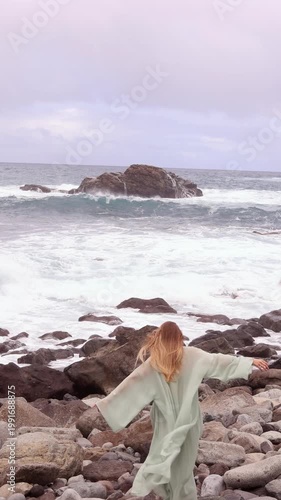 A vertical full-length shot from behind of a blonde woman in a flowing mint-green outfit walking over large coastal rocks toward powerful ocean waves crashing against a big rock under a moody sky.