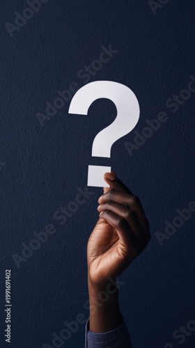 Dark-skinned hand holding white paper question mark against deep navy textured wall — minimal modern interior, warm window light