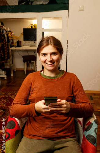 Happy young woman sitting in a colorful armchair and using a smartphone at home. Smiling female scrolling social media or texting on mobile phone in a cozy interior. Gen z girl