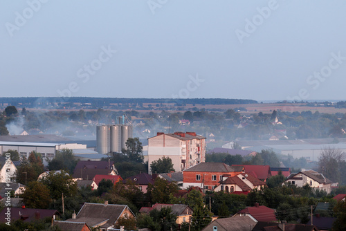 Air pollution from industrial factory pipes over a small town. Concept of environmental issues, smog, and carbon emissions affecting residential areas. Grain elevator