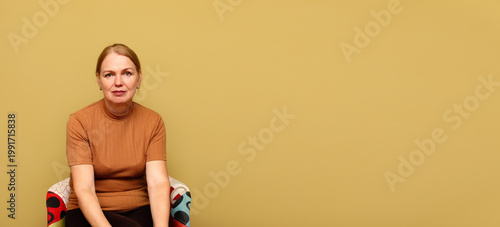 Middle-aged woman sitting in a colorful armchair against a solid yellow background with large copy space. Portrait of a calm female in a brown t-shirt. Minimalist studio photography for advertising