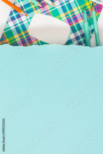 Creative back to school background with colorful plaid notebook and stationery items under a torn light blue paper edge. Minimalist vertical flat lay with large copy space for text and design.