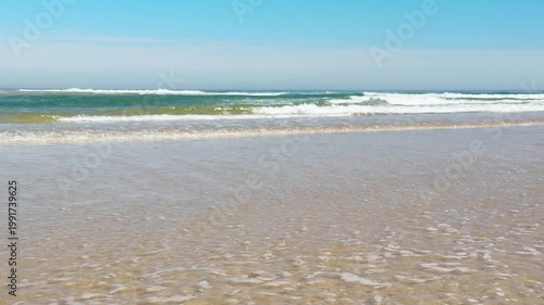 Serene ocean waves gently washing over a vast sandy shore under a clear blue sky. A tranquil and peaceful coastal landscape showing the tide receding from the wet, golden sand