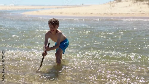 Little boy in blue swim trunks having fun and playing with a big wooden stick in the shallow water of the sea on a bright, sunny day, enjoying his summer vacation by the ocean shoreline