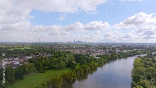 A wide aerial view of the Main River winding through a lush green landscape. In the background, the prominent skyline of Frankfurt am Main is visible under a slightly cloudy summer sky.