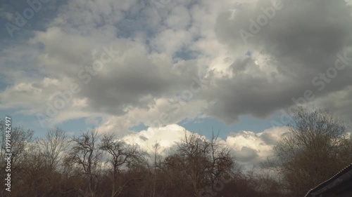 Dramatic light and dark clouds drift over a horizon of leafless trees. This calm atmospheric scene features soft daylight and natural textures with a farm building roof seen in the corner. 