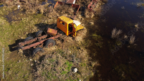 aerial shot of an old, rusted orange and white heavy-duty truck cabin and chassis abandoned in a swampy, waterlogged field