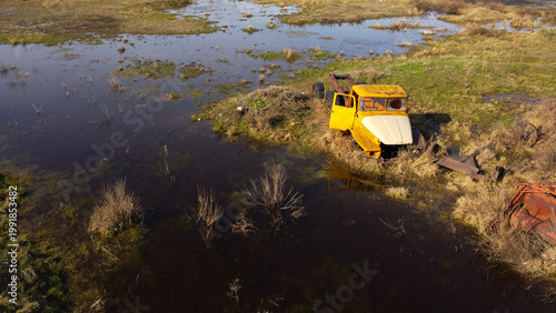 aerial shot of an old, rusted orange and white heavy-duty truck cabin and chassis abandoned in a swampy, waterlogged field