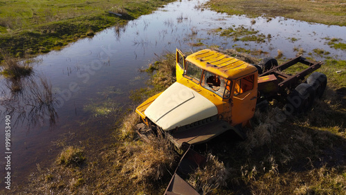 aerial shot of an old, rusted orange and white heavy-duty truck cabin and chassis abandoned in a swampy, waterlogged field