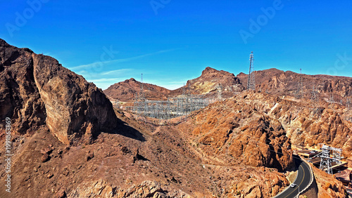 Desert Power Grid Near Hoover Dam. Rocky desert landscape near Hoover Dam with transmission towers, electrical infrastructure, winding road, and clear blue sky in the American Southwest.
